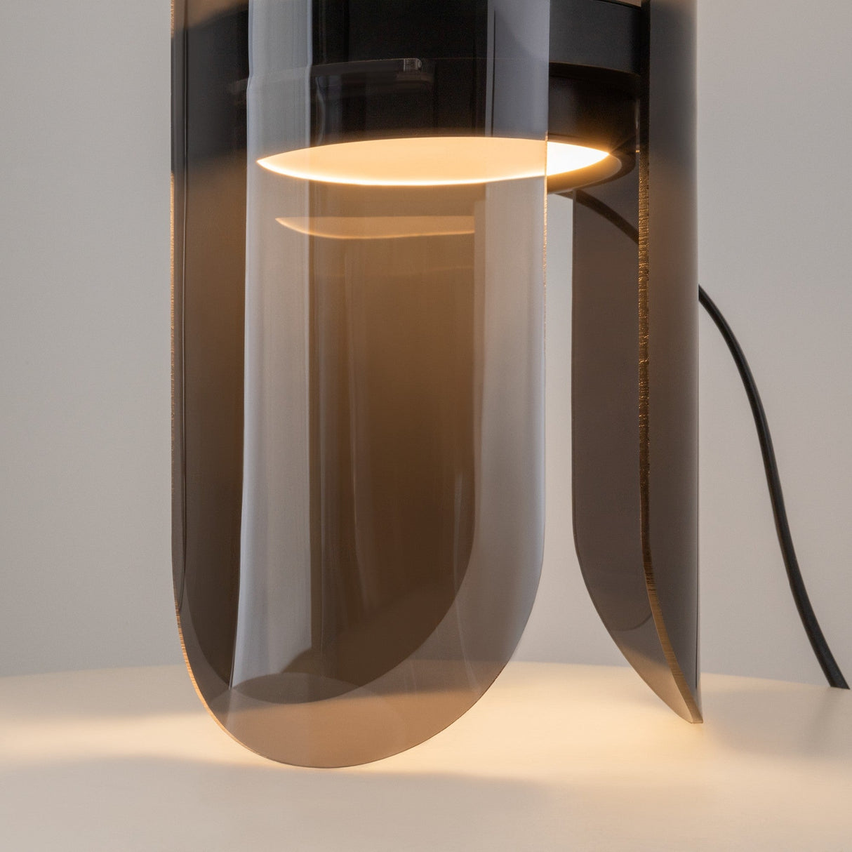 The Insight LED Table Lamp - Black has a minimalist cylindrical design with a translucent shade. Its warm ambient light diffuses through a clear dark-toned cover, casting a soft glow on the white surface below. A visible black power cord adds to its modern touch.