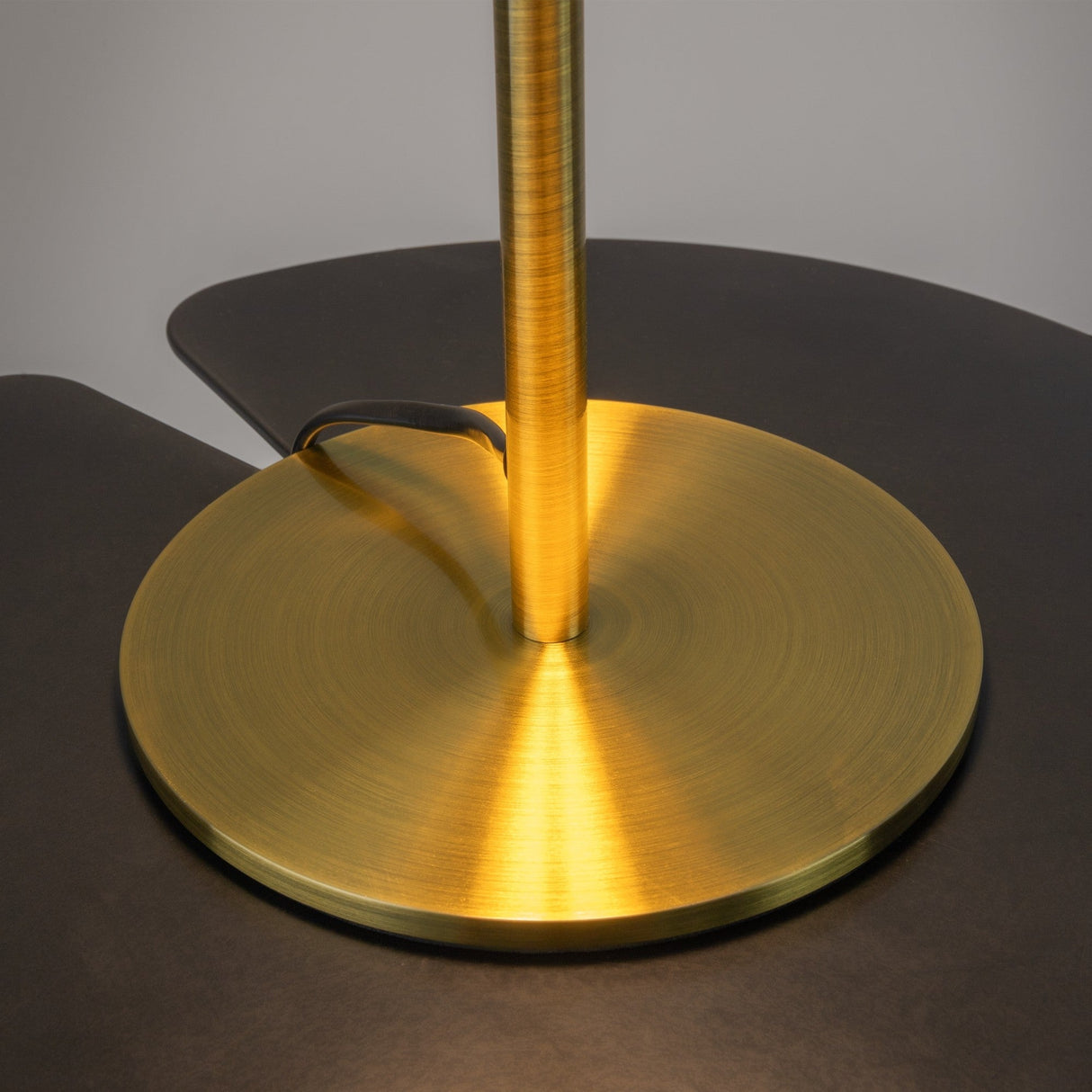 A close-up of the Mystic LED Table Lamp - Brass reveals a golden circular base and slim vertical stem. Its brushed metal texture and brass finish suit the spherical glass shade for a warm glow. A black cord is partially visible, contrasting with the shiny surface.