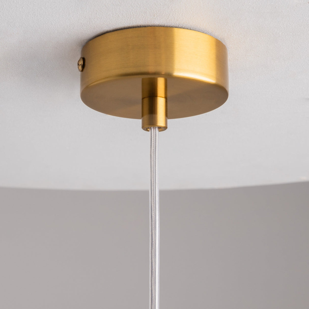 Close-up of the minimalistic and modern Flusso Long Chain Pendant Light - Bronze With Clear Glass Ball, featuring a brass ceiling fixture and round canopy. A clear cord with bronze-finished chains descends against a white ceiling and gray wall.
