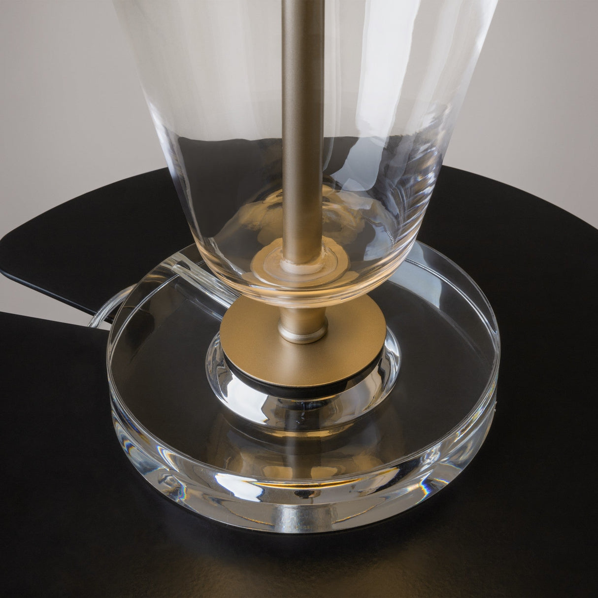 Close-up of the Verre Table Lamp featuring a brass rod on a black surface, with a clear glass base and white linen shade. Its sleek, minimal design offers a perfect blend of glass and metal elements.