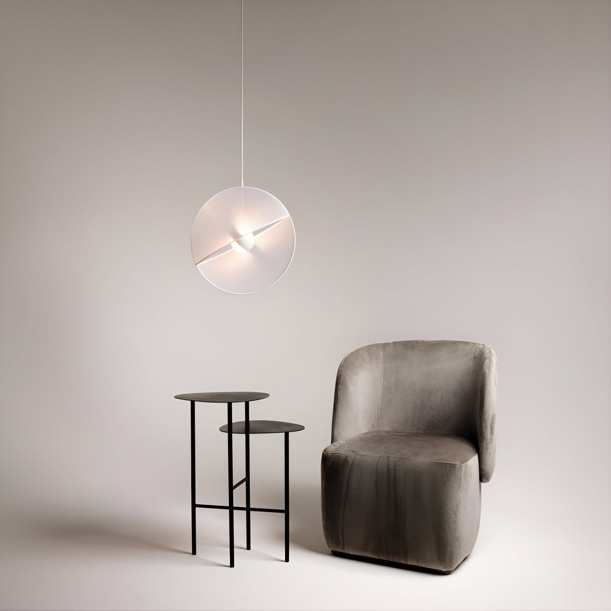 A minimalist interior features a soft taupe armchair, two black nested round tables, and a Reflex 1 Light LED Disc Pendant in white with an adjustable wire casting a warm glow against a plain beige wall.