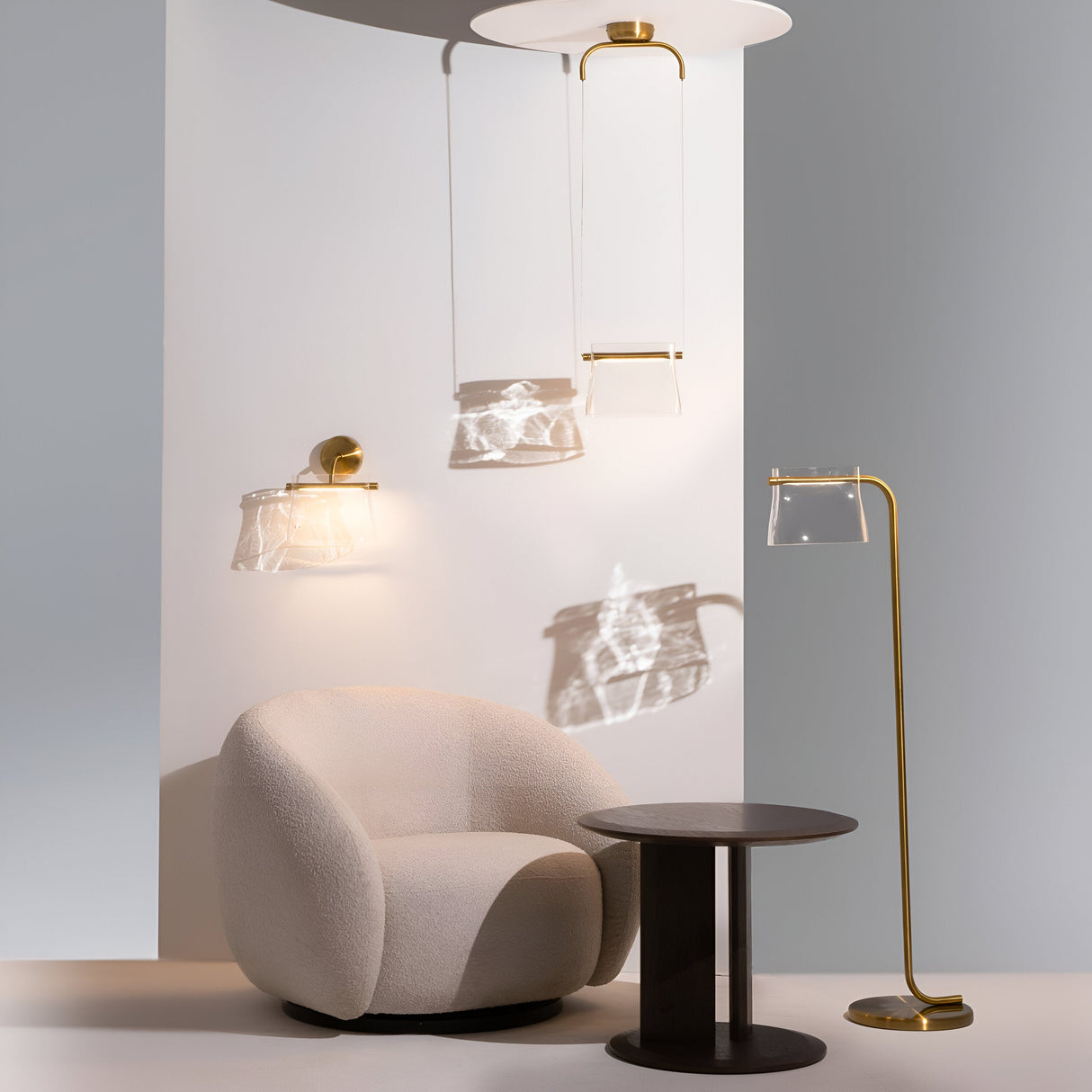 A modern interior includes a plush beige armchair, a sleek round dark wooden side table, a floor lamp with a clear lampshade, and the Cabaret LED Pendant Light in brass. This lighting fixture casts artistic shadows on the light gray wall.