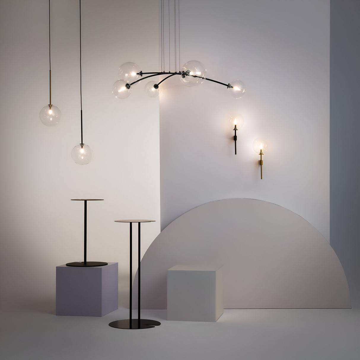 A minimalist interior embodies modern elegance with contemporary suspension lights featuring globe bulbs. Two slender tables rest on square platforms, while a semi-circular wall detail and the Mood 6 Light Suspension - Black With Clear Glass enhance the design.