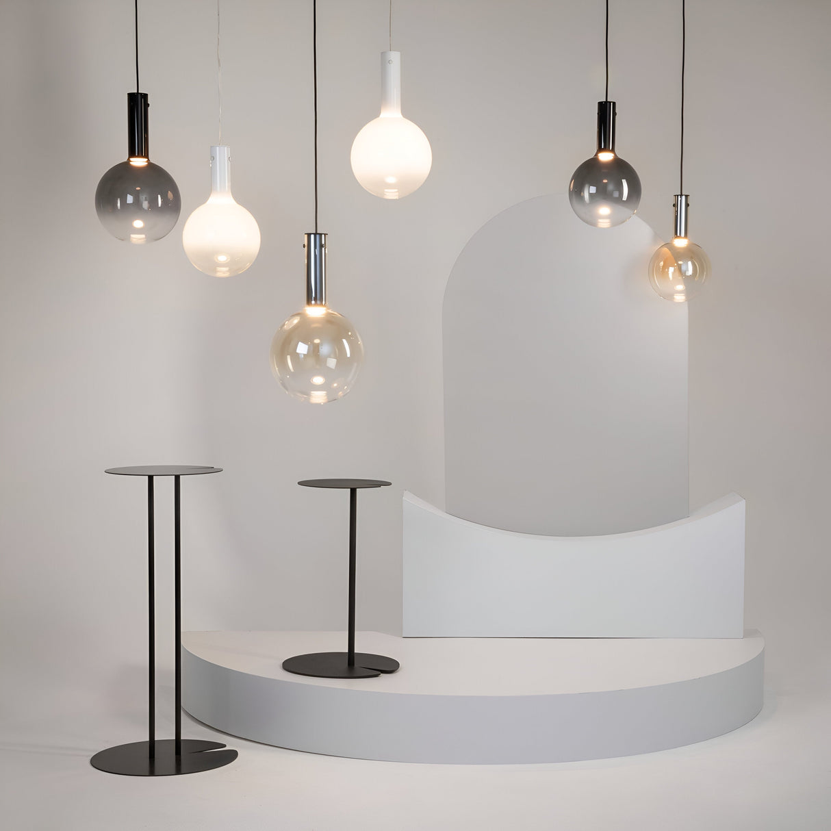A minimalist setup features misty white glass spheres and Nebula LED Pendant Lights in black and translucent hues, suspended above two slender black round tables and a white curved platform against a neutral backdrop.