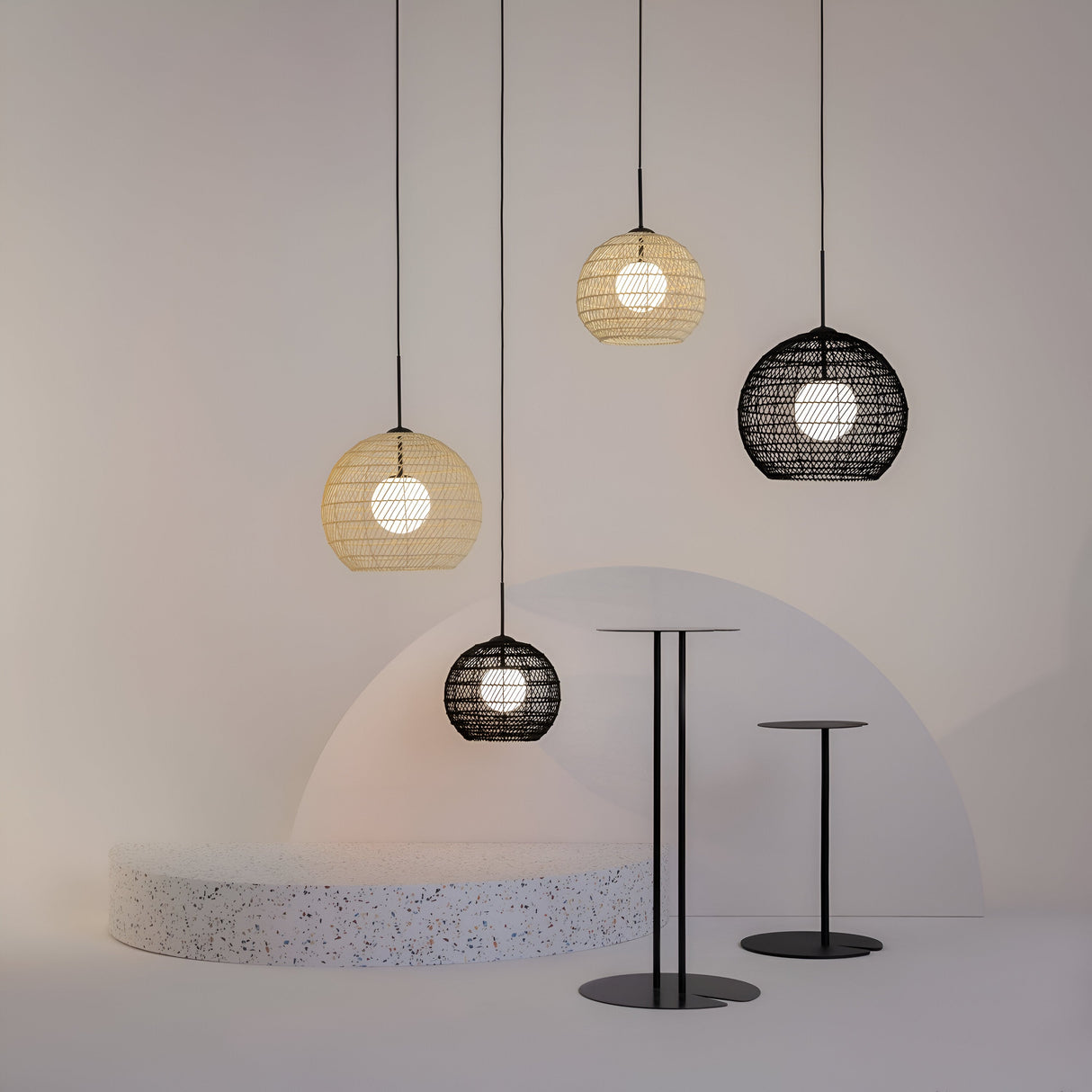 A modern room showcases four Cane Pendant Lights (40cm, Black Wicker with White Glass Diffuser) casting a warm glow. Below are two tall round pedestal tables of varied heights. A terrazzo semi-circle platform graces the floor against a light wall, enhancing the organic design aesthetic.