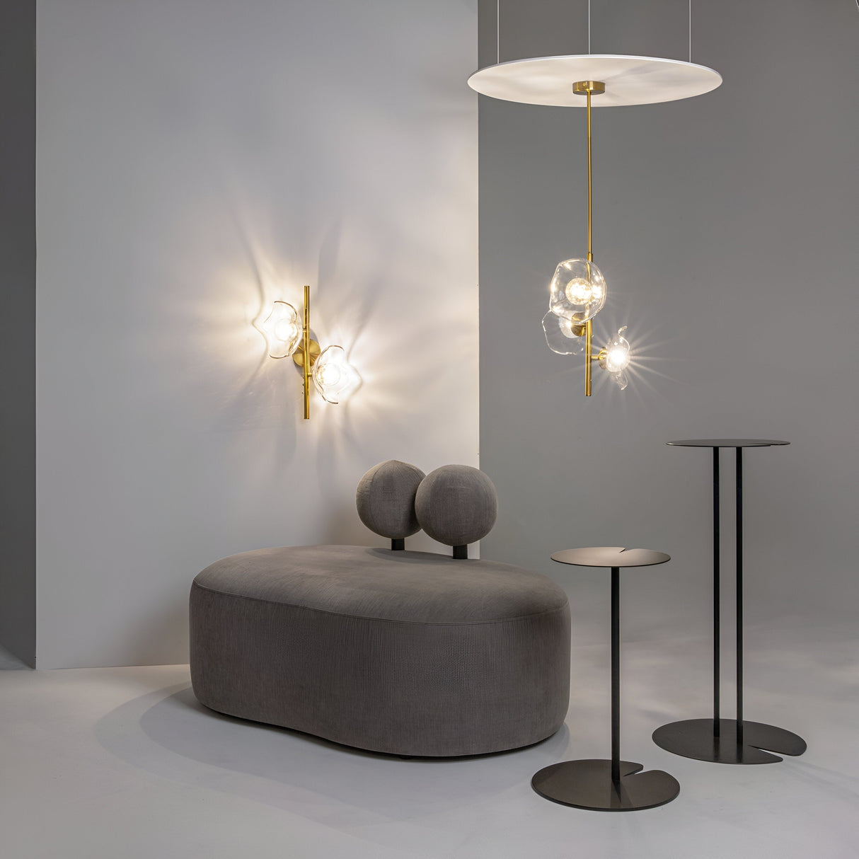 Modern interior with a gray cushioned bench, round cushions, two black side tables, and stylish Art Deco accents. The Miracle 3 Light Pendant in brass has clear glass shades and spherical bulbs that emit a soft glow against the light gray wall.
