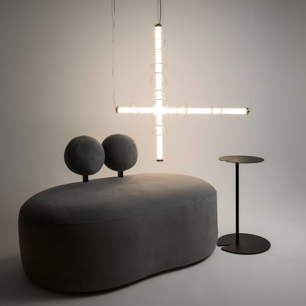 A modern interior features an oval gray sofa with two spherical cushions and a vertical black side table. Overhead, a Cosmo LED Linear Suspension Light in black with clear glass enhances the minimalist aesthetic, casting a futuristic glow on the neutral background.
