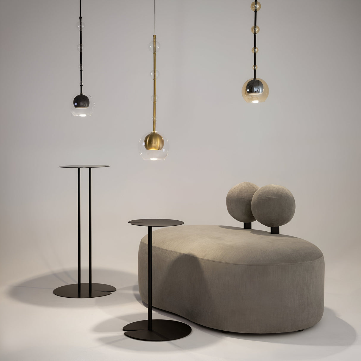 A modern interior showcases a gray oval sofa with two round backrests, flanked by black minimalist side tables with round tops. The scene is illuminated by three Ros 1 light pendants featuring amber glass shades and black finishes, enhancing the neutral color palette.