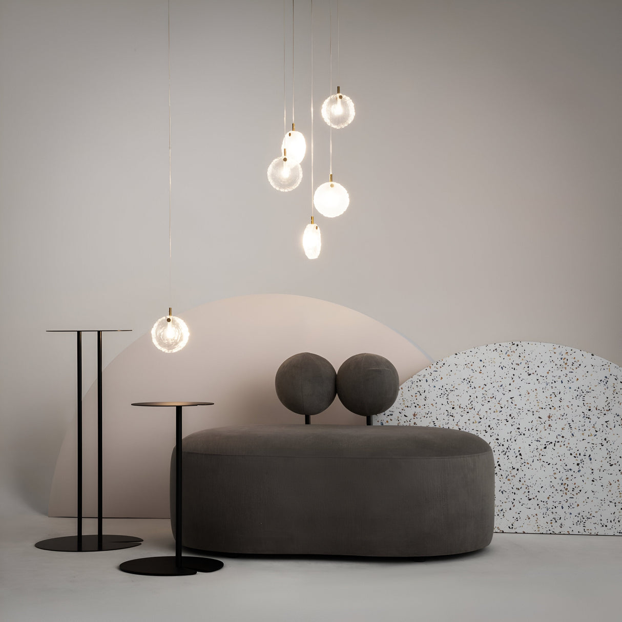 A minimalist room with a gray modern sofa and twin black side tables showcases a Frozen 5 Light Cluster Pendant - Brass, featuring cast solid glass lampshades. The brass fixtures hanging lights are complemented by abstract beige and speckled arches to enhance the contemporary aesthetic.
