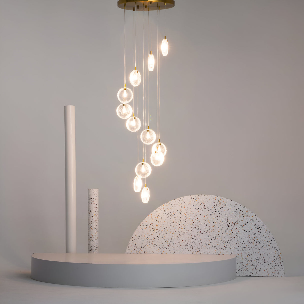 A modern art installation features a cluster of glowing pendant lights above a minimalist stage, enhanced by the elegance of Frozen 12 Light Spiral Pendant - Brass With Wood Cut Clear Cast Glass. The stage has circular shapes in neutral tones, speckled patterns, and a plain gray backdrop.