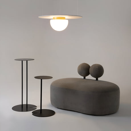 A modern room features a minimalist design with two round black side tables of varying heights, an oval gray sofa with rounded back cushions, and the Glassy Ceiling Flush Light - Gold With White Matte Glass. A plain, light background complements this elegant aesthetic.