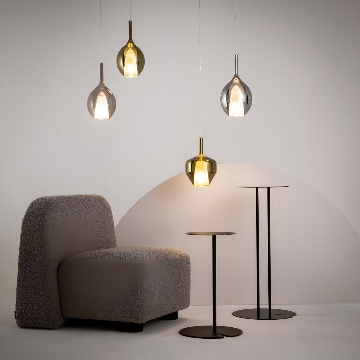 A modern industrial design features a textured gray chair and four Duality Round Pendant Lights with black frames and mirrored gold glass shades. Two sleek, round side tables with thin metal stems complement the space, while the light gray walls partial circular design enhances the vibe.