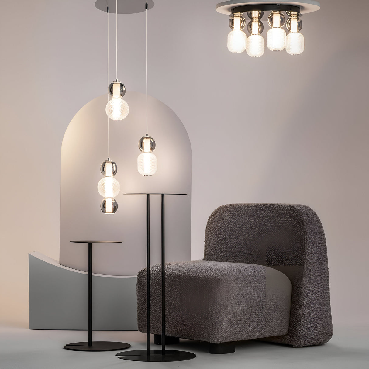 A modern interior features a plush gray chair, two sleek black side tables, and the Drop LED Cluster Pendant Light 33W 3000K - Chrome descending from the ceiling against a neutral arch-shaped backdrop. The minimalist setting elegantly blends style with energy-efficient lighting.