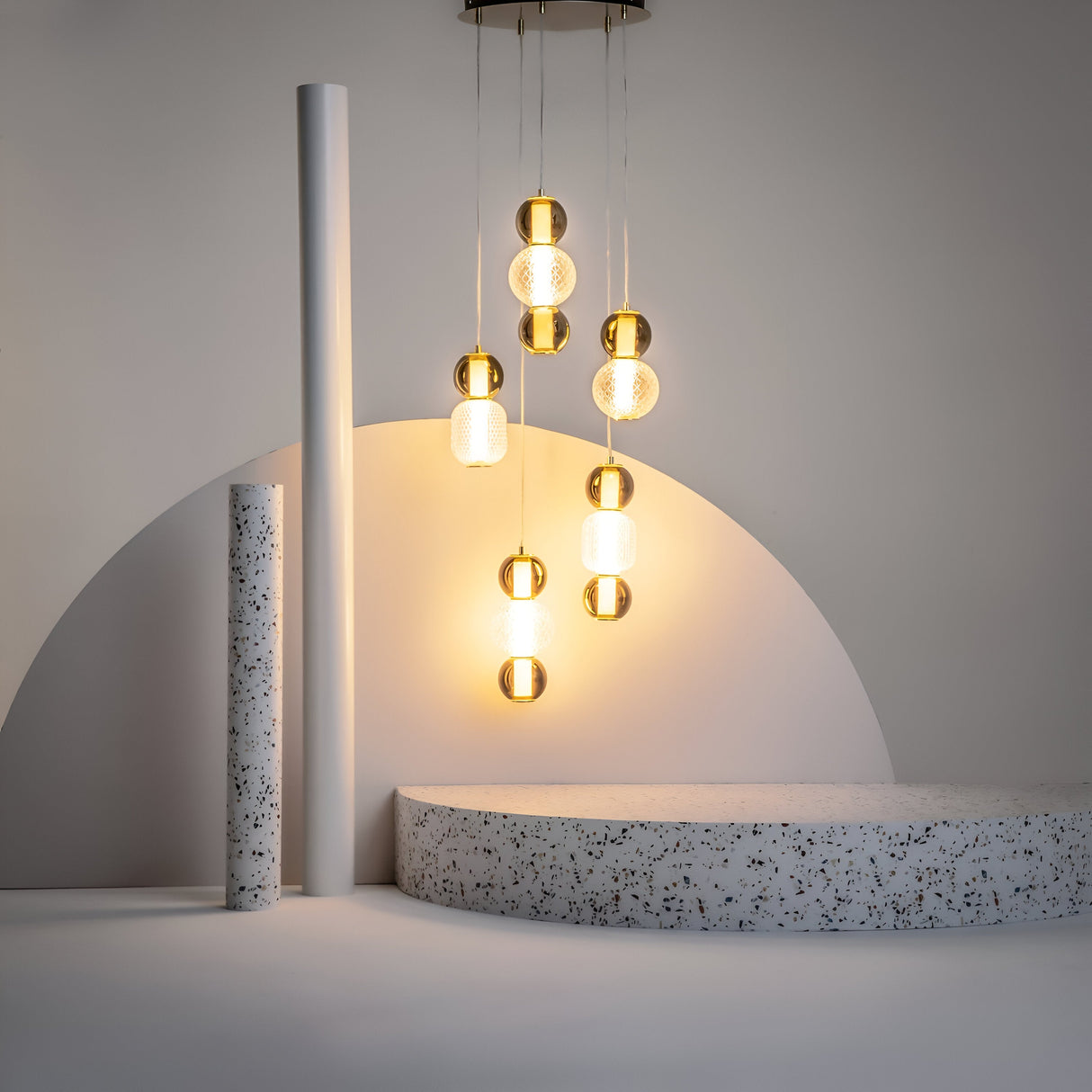 A modern indoor scene features the Drop LED Cluster Pendant Light 80W 3000K - Gold, highlighting a white speckled terrazzo semicircular platform. Beside it, a vertical pipe against a neutral background enhances the energy efficiency and contemporary elegance of the space.