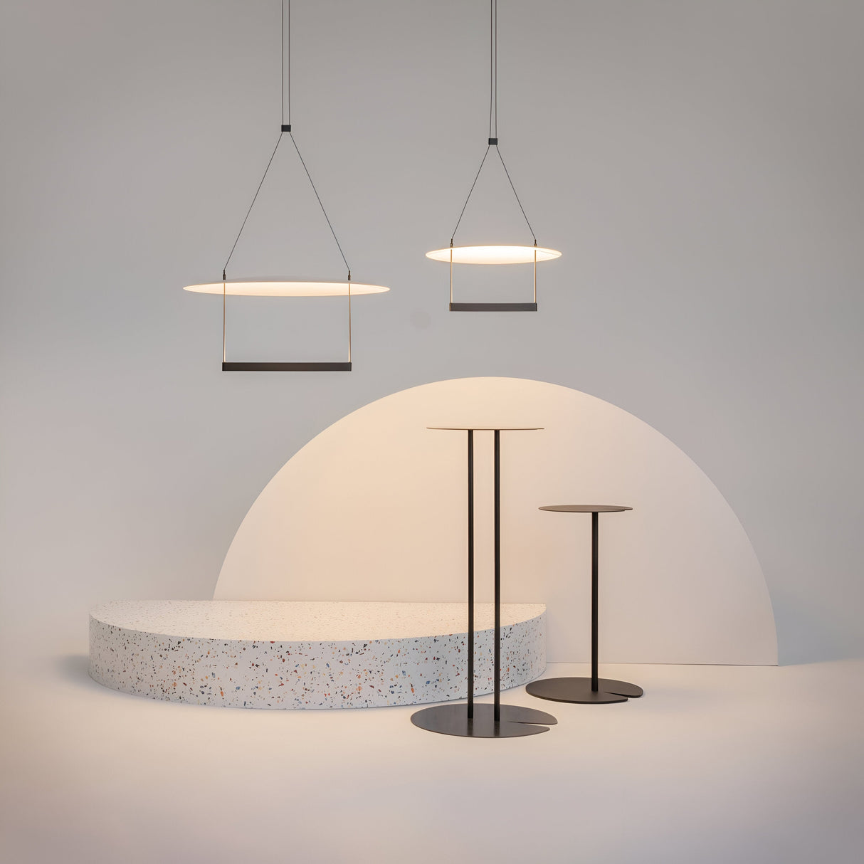 A minimalist interior with modern elegance features energy-efficient Ambience LED Round Flat Small Pendant Lights in black above a terrazzo oval table. Two slender black bar tables stand to the right, set against a white backdrop with a semi-circular wall accent.