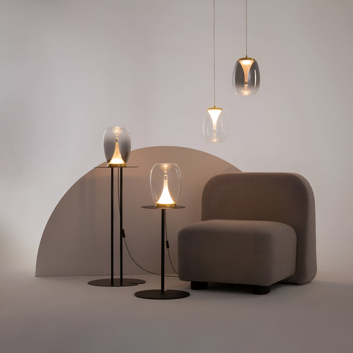 A cozy scene with a plush grey armchair beside a semi-circular backdrop. Three Splash LED Pendant Lights, 3000K 7W in gold with clear glass, hang above. Two matching floor lamps with glowing bulbs stand on sleek gold metal stands next to the chair.