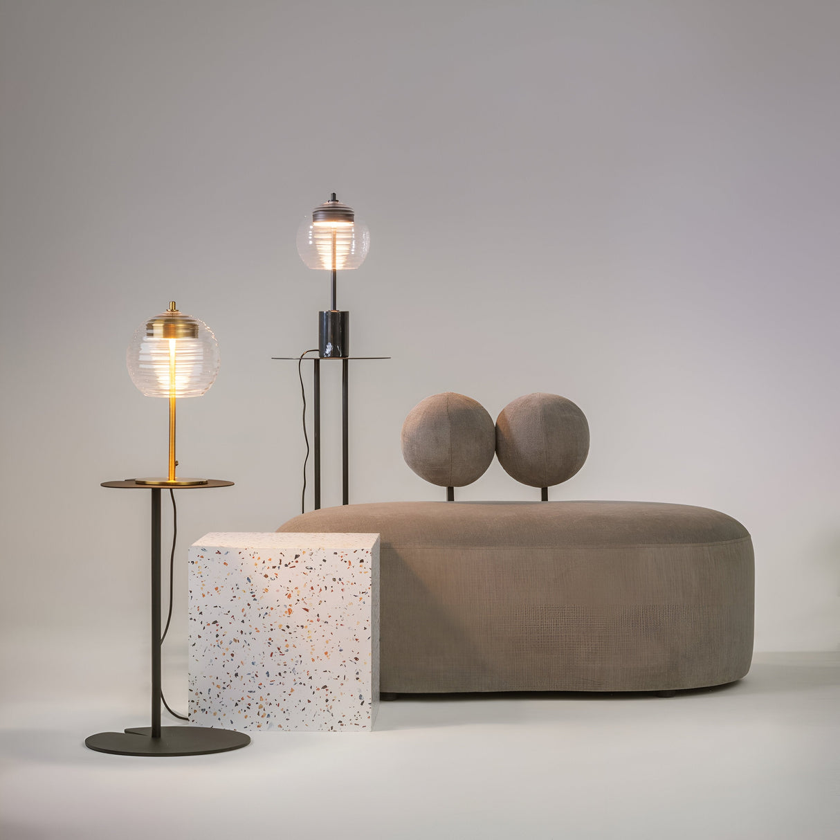 A modern setting features a gray sofa with round backrests, two Mystic LED Table Lamps in brass with spherical glass shades, and a white cube speckled in multiple colors against a light gray wall.