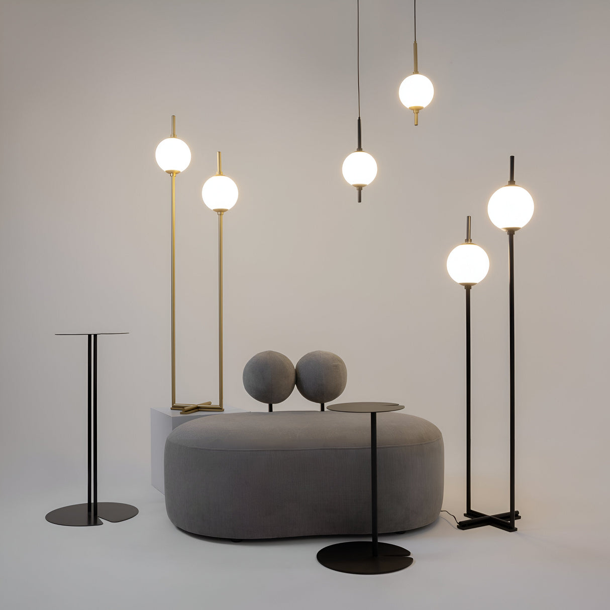 A minimalist room showcases a modern gray oval sofa with circular cushions, flanked by standing lamps and The Sixth Sense LED Pendant Light featuring white glass spherical shades. Two small round side tables complete the contemporary decor, highlighting the minimalist design.