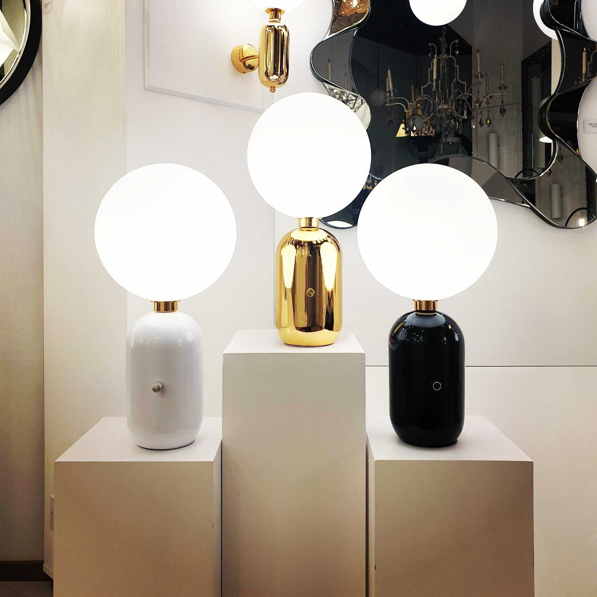 Three Niori Pallo Table Lamps with opal glass globes on white, gold, and black capsule bases, displayed on white plinths near mirrors and a chandelier.