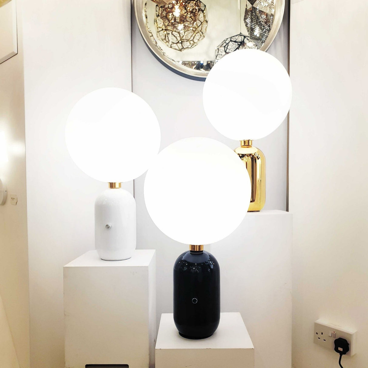 Niori Pallo Table Lamp 14cm in gold with opal glass globe shade displayed on a white pedestal beside white and black versions, with a round mirror above.