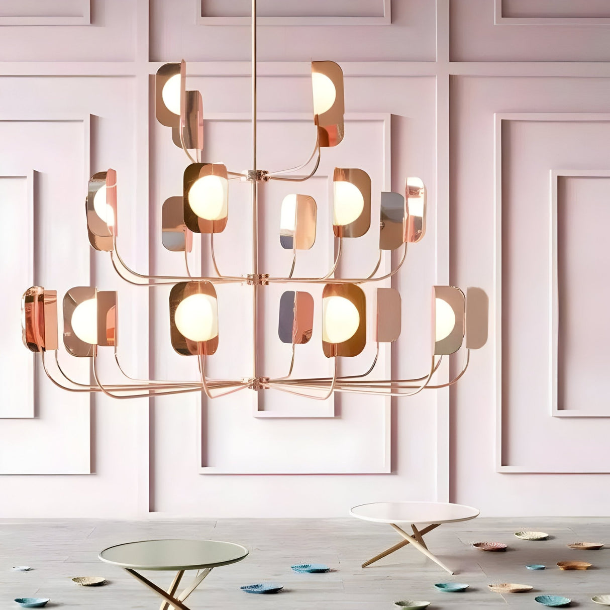 The Petal 12 Light Chandelier - Copper hangs before a pink paneled wall, its rose-gold arms and round bulbs adding nature-inspired elegance above two round tables with gold legs and colorful coasters.