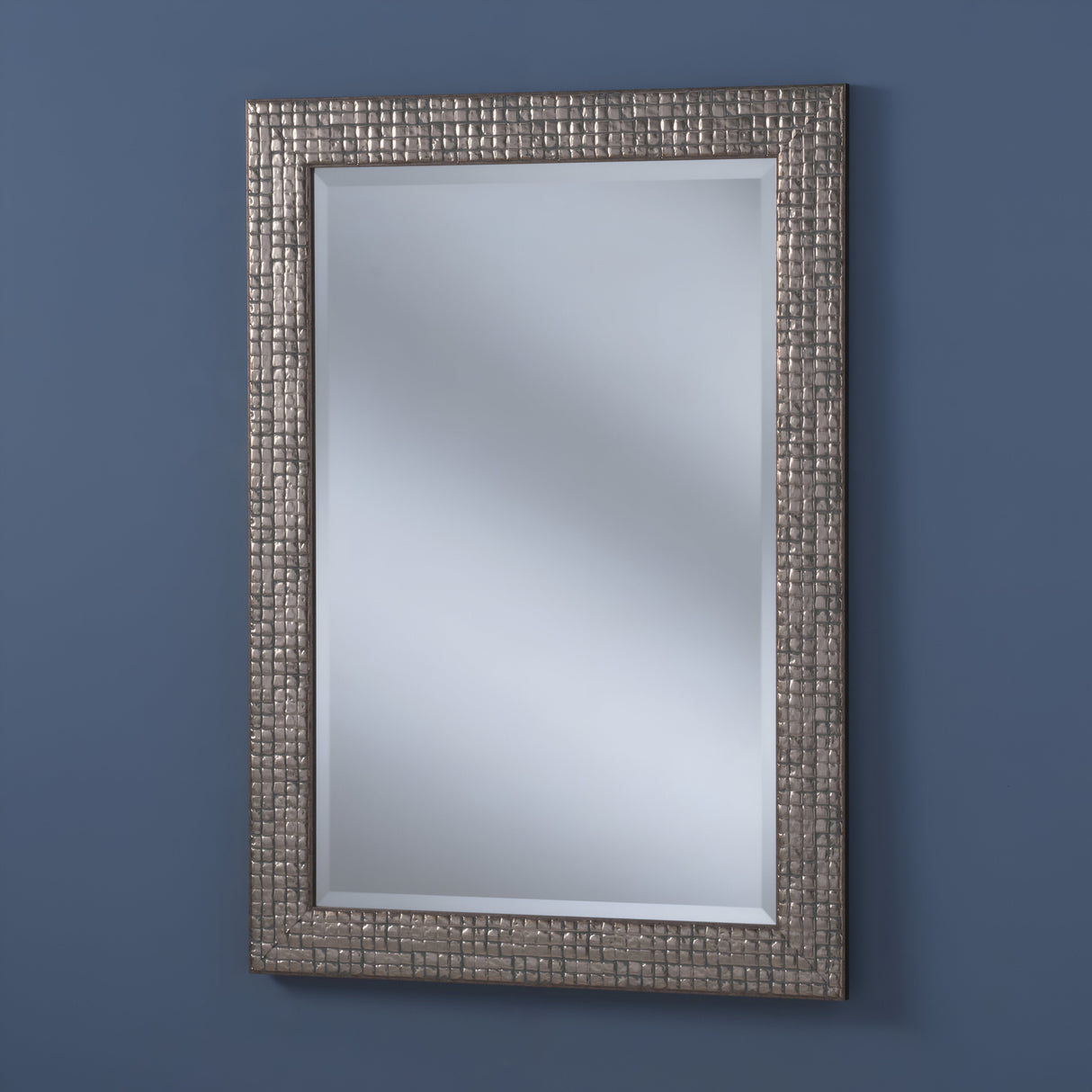 The Iridessa Rectangle Wall Mirror - Antique Silver showcases a textured, metallic frame with a grid pattern, offering vintage-inspired elegance and a clear reflective surface on a solid blue wall.