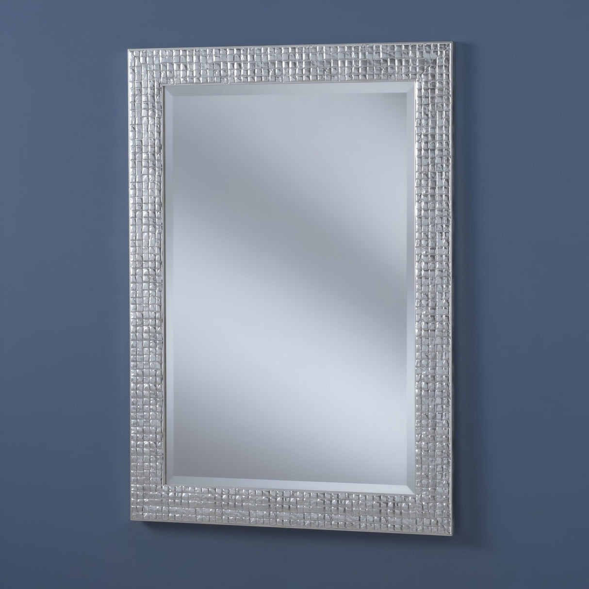 The Iridessa Rectangle Wall Mirror - Silver is a stunning home decor piece with a textured silver frame mounted on a blue wall. It subtly reflects light and boasts an elegant grid-like pattern, enhancing its simple design.