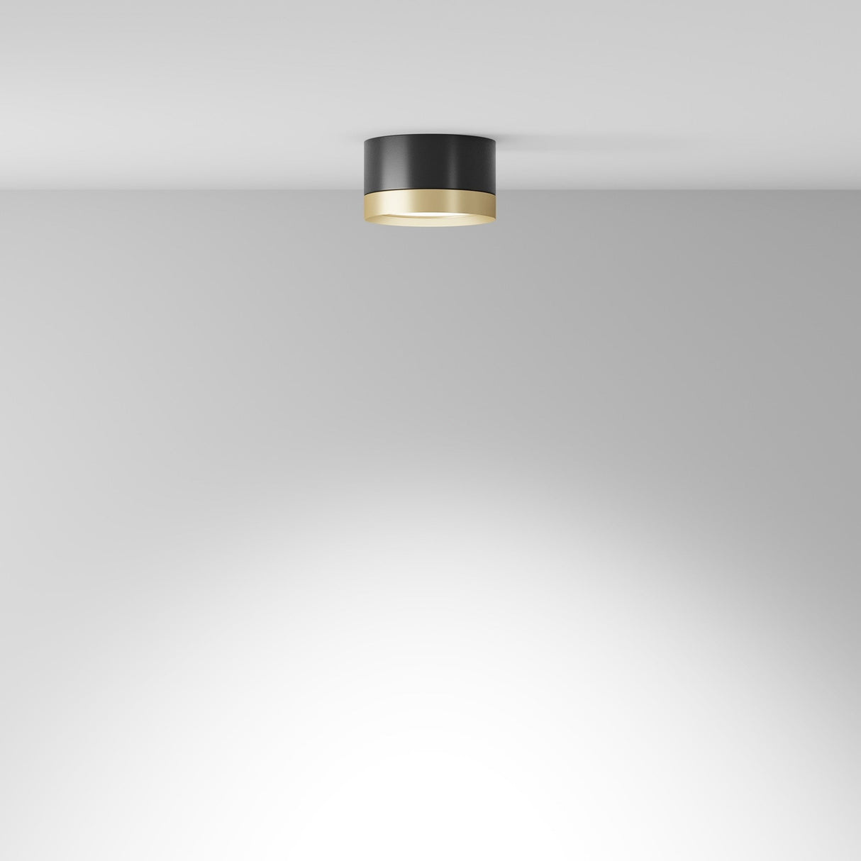 The Hoop Small Round Surface Downlight in black with a gold rim is mounted on a white ceiling, casting a soft glow on the plain wall below and effortlessly blending into contemporary interiors.