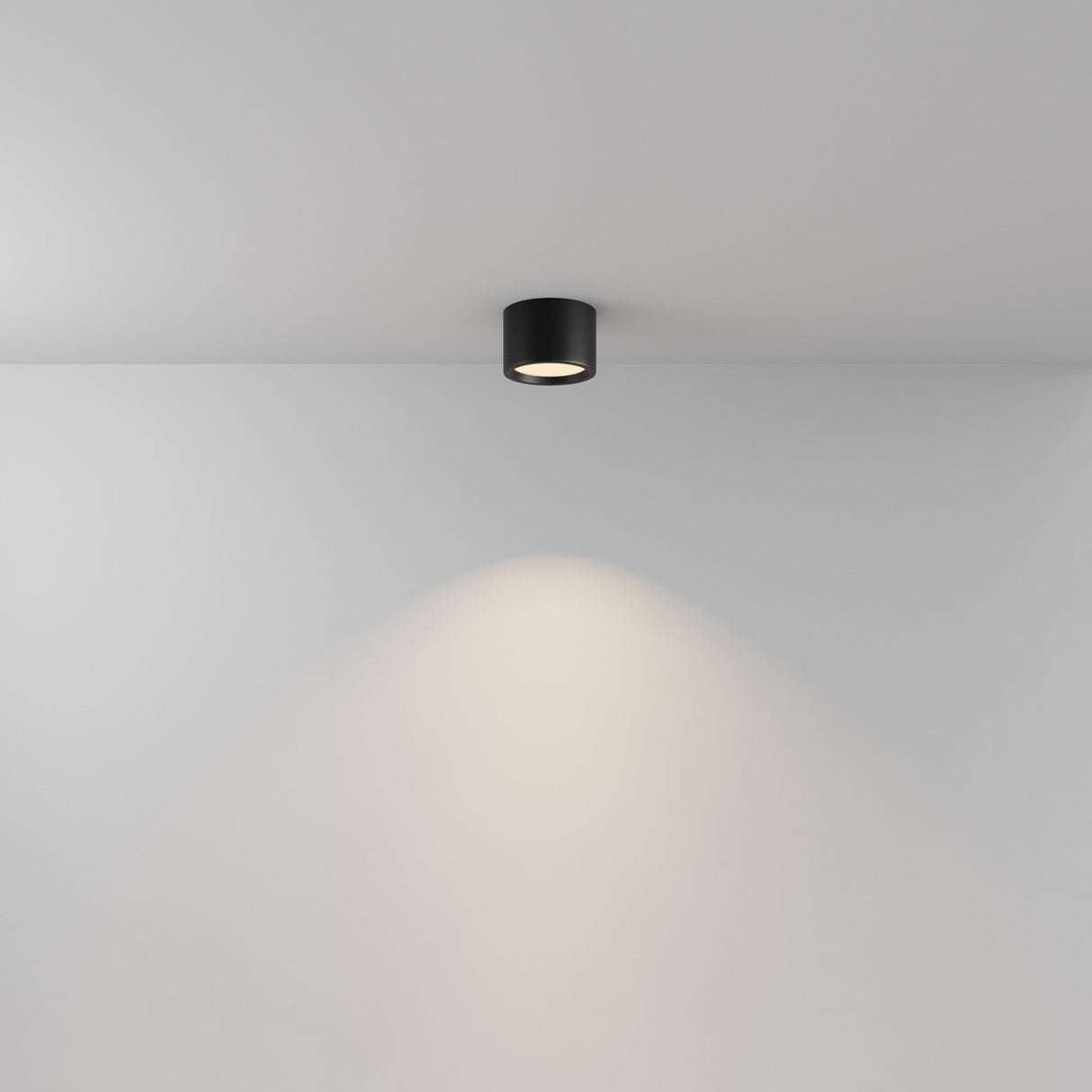 The Hydra LED Round Surface Downlight 10W 3000K in black casts a soft, circular glow on a white wall, creating a warm and inviting ambiance with its sleek cylindrical design and energy-efficient LED technology.
