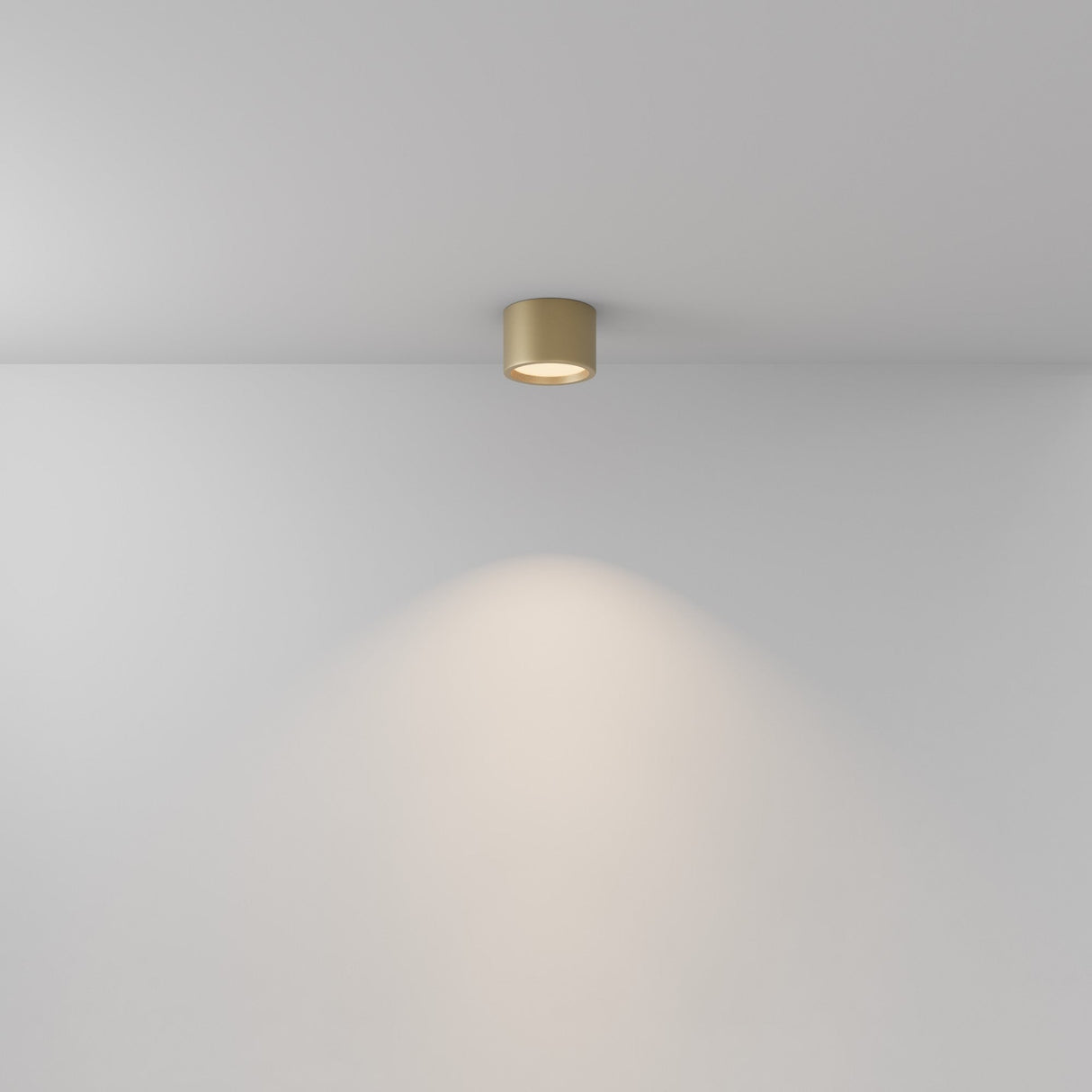 A minimalist scene includes a white ceiling and wall highlighted by the Hydra LED Round Surface Downlight 10W 3000K in matte gold. Its cylindrical design and IP44 rating cast a soft, circular glow, enhancing the elegant ambiance.