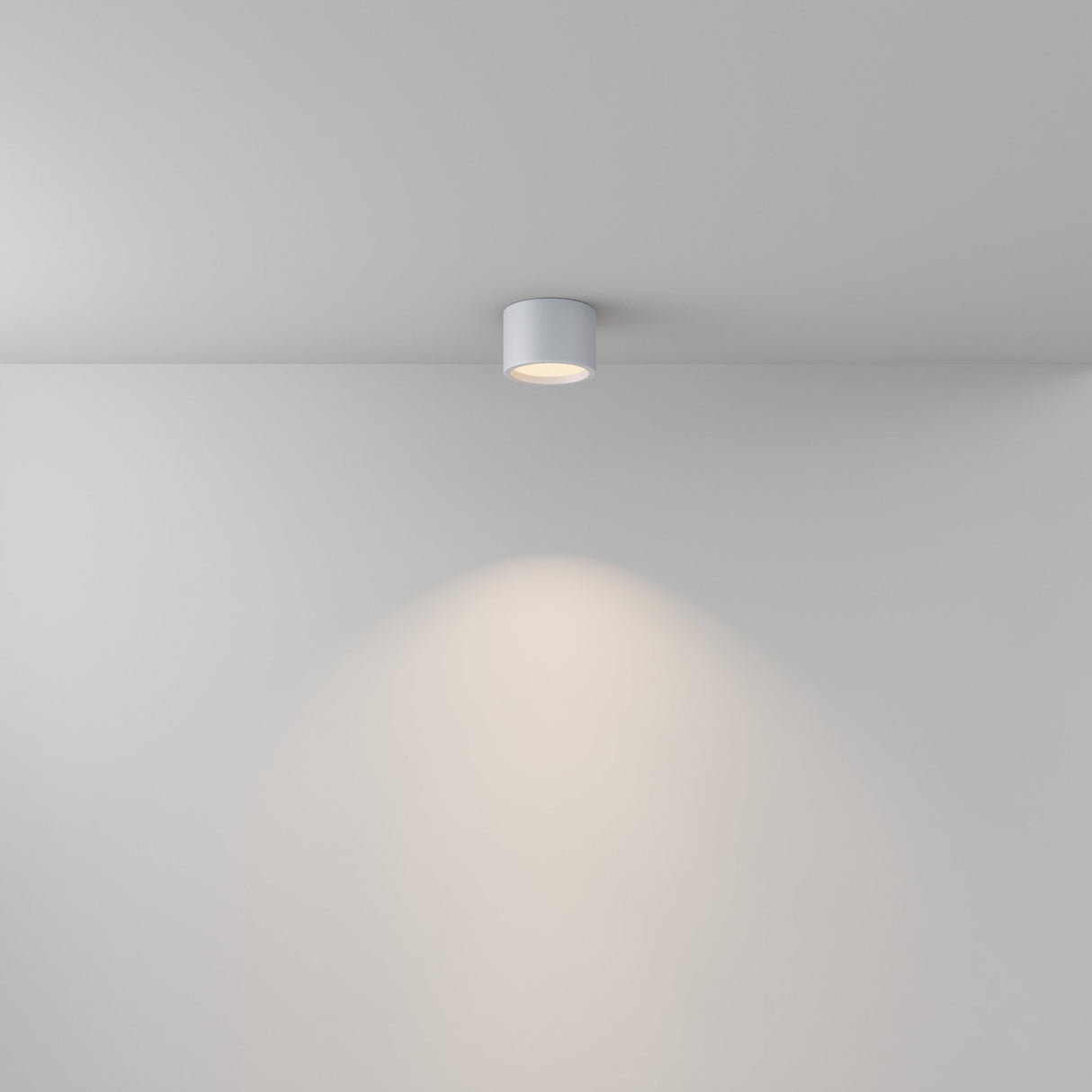 The Hydra LED Round Surface Downlight 10W 3000K - White, rated IP44, is mounted on a white ceiling. It emits a soft, focused glow directly below, creating a subtle light cone on the wall and maintaining a minimalistic and clean setting.