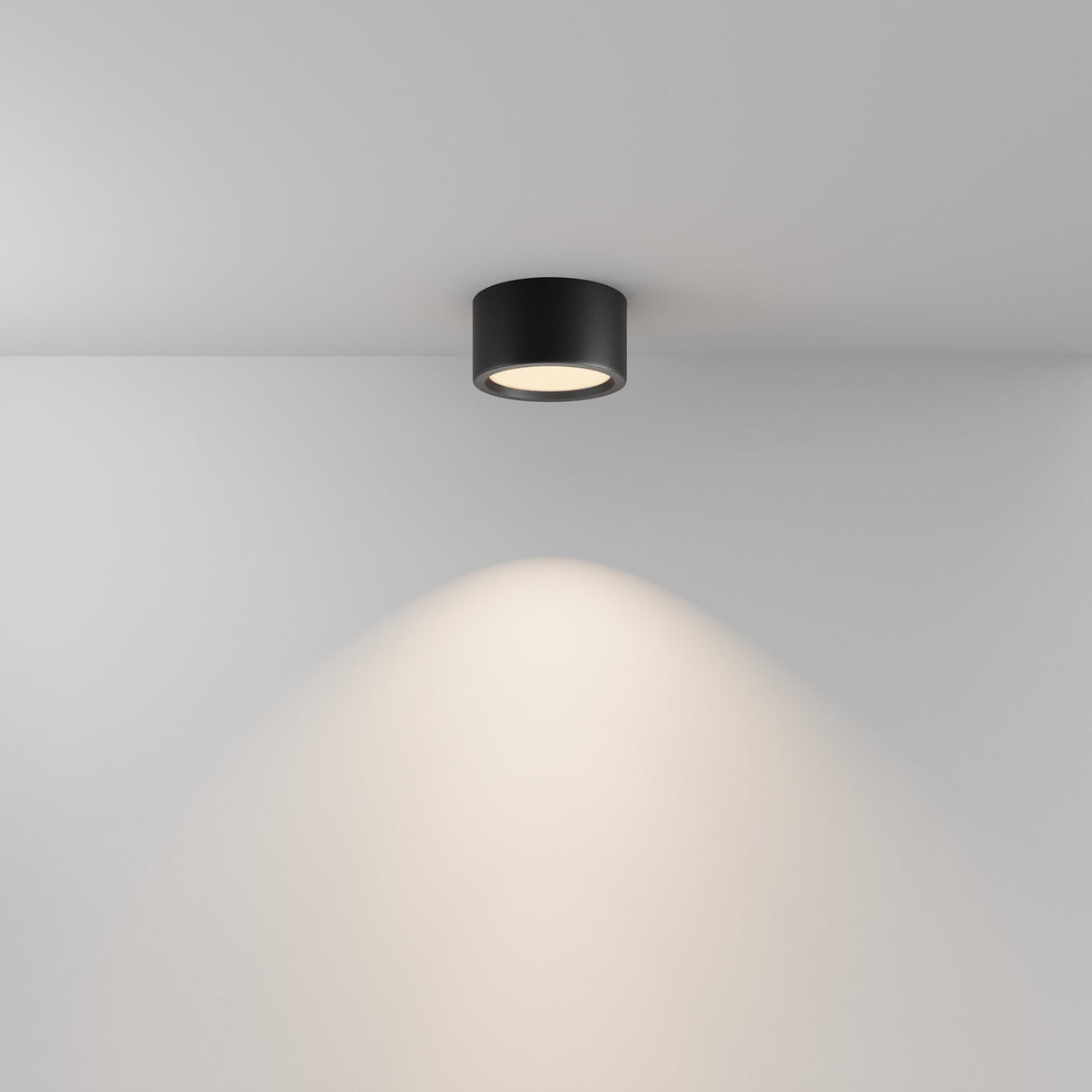 The Hydra LED Round Surface Downlight 15W 3000K in black is mounted on a white ceiling, providing a soft, warm glow on the wall below.