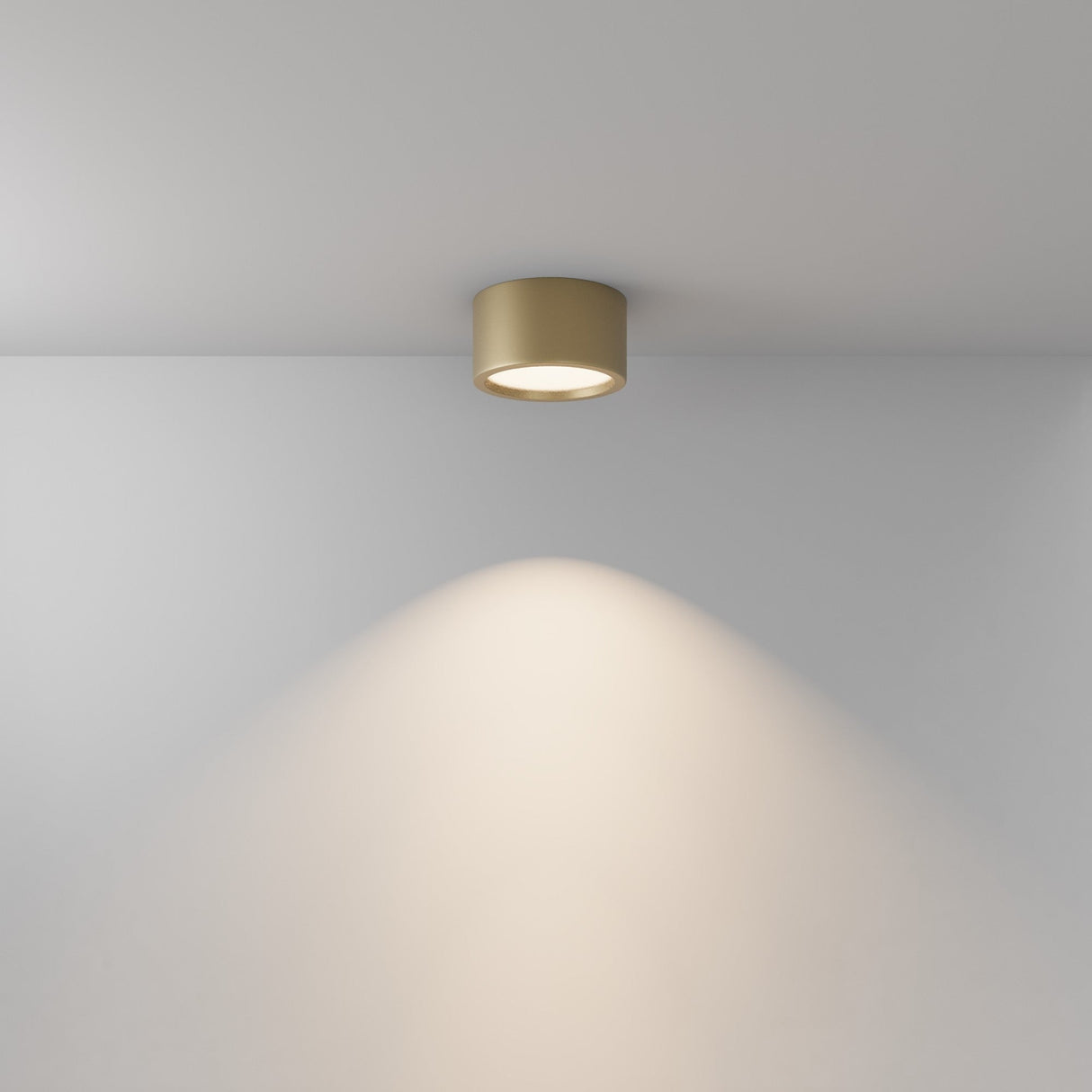 The Hydra LED Round Surface Downlight 15W 3000K in Matte Gold is mounted on a white ceiling, casting a warm, focused light with a cone-shaped illumination pattern onto the wall.