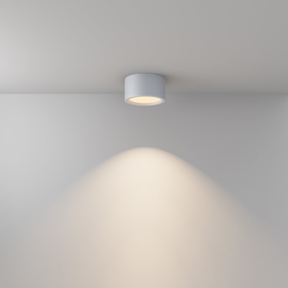 The Hydra LED Round Surface Downlight 15W 3000K, with its IP44 rating, casts a soft, circular glow on a white wall and ceiling. This cylindrical, white LED fixture seamlessly blends into any modern setting.