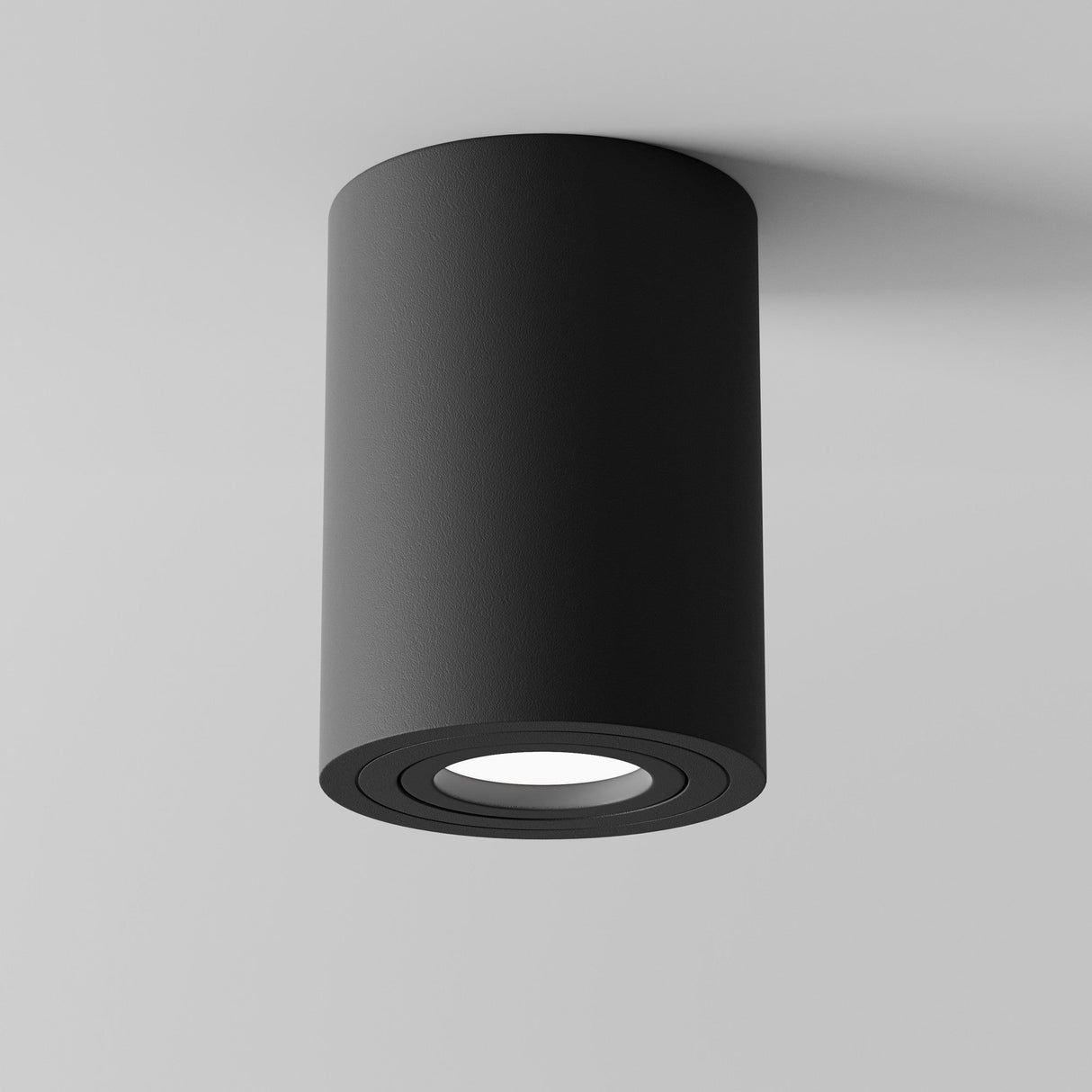 The Atom Round Surface Mounted Downlight in black offers ambient lighting with concentric details around its downlight opening, all set against a smooth white ceiling. Its modern cylindrical design and matte finish highlight its minimalist appeal.