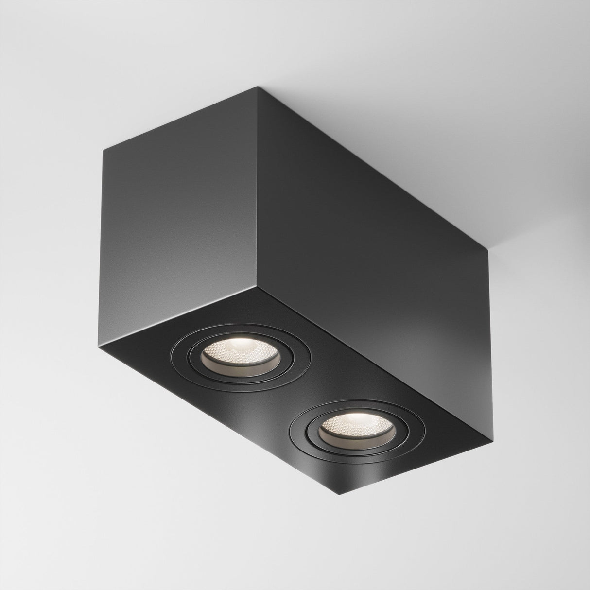 The Atom Twin Adjustable Rectangular Surface Mounted Downlight in black features a modern minimalist design with two recessed circular lights and an adjustable downlight. This flush-mounted fixture emits soft white light, enhancing the aesthetic of a white ceiling.