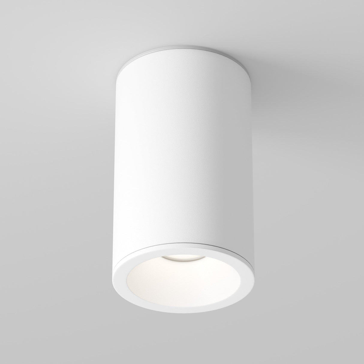 A modern, cylindrical Zoom Small Round Surface Downlight in white is mounted on a light gray ceiling. It emits a soft, warm glow from its round opening, providing a minimalist look with an IP65 rating for enhanced durability.