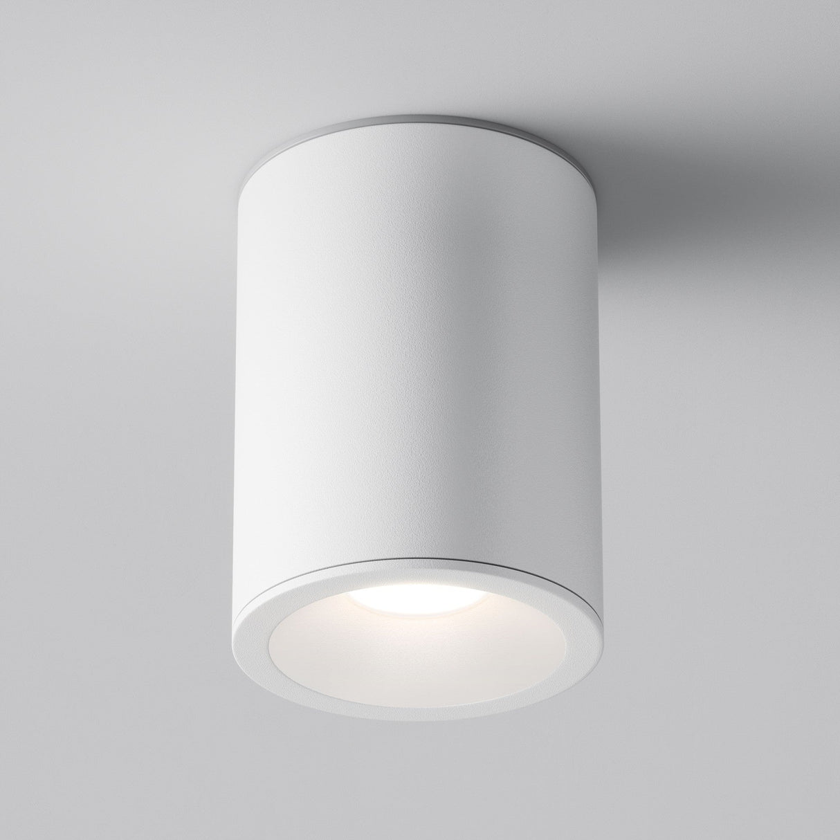 The Zoom Round Surface Downlight - White is a sleek, cylindrical fixture in matte white, mounted on a ceiling. Made with an aluminium body and IP65 protection, it emits a soft glow with its smooth, minimalist design.