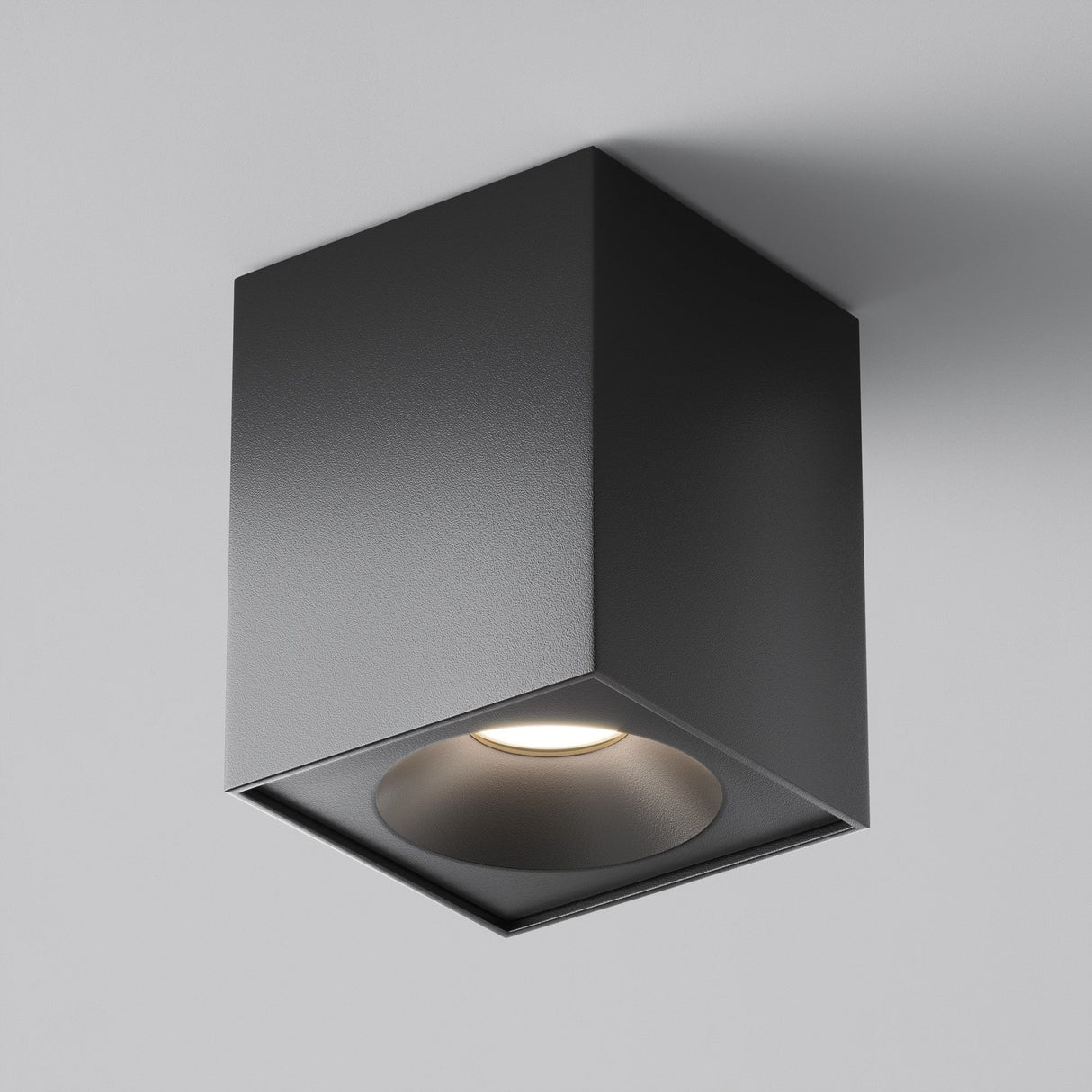 The Zoom Square Surface Downlight - Black is a ceiling-mounted light with a matte black finish and minimalist design. It emits a soft glow from its recessed bulb, offering a modern, sleek look against a white ceiling.
