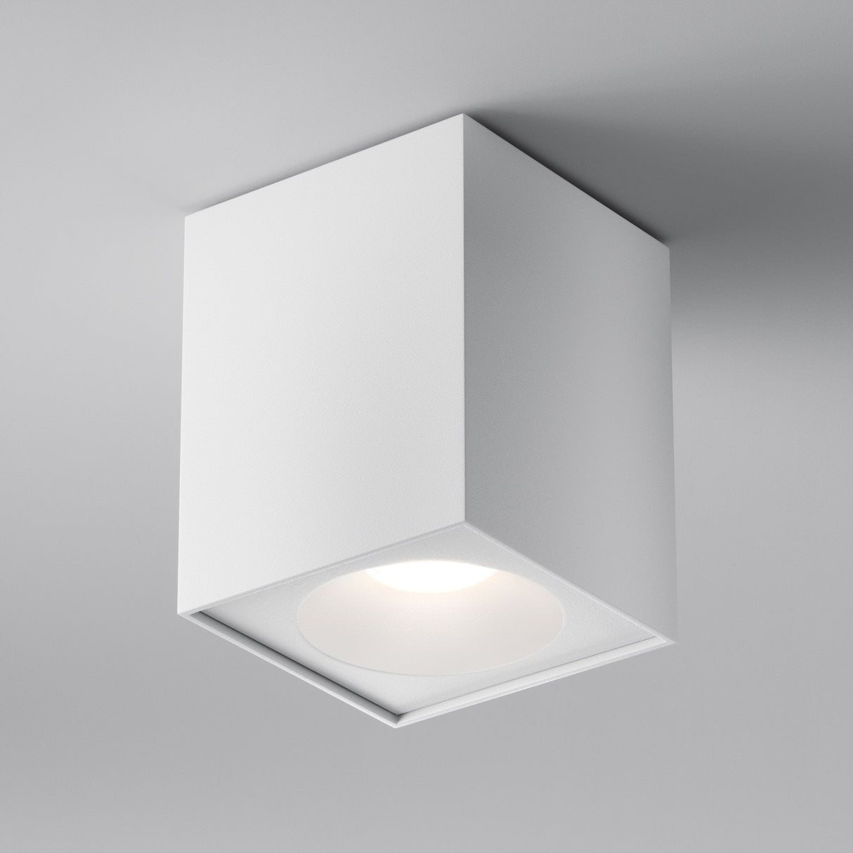 The white Zoom Square Surface Downlight, with its cube shape, is mounted on a gray ceiling. It features an IP65 rating and provides a soft glow, ideal for modern interiors.
