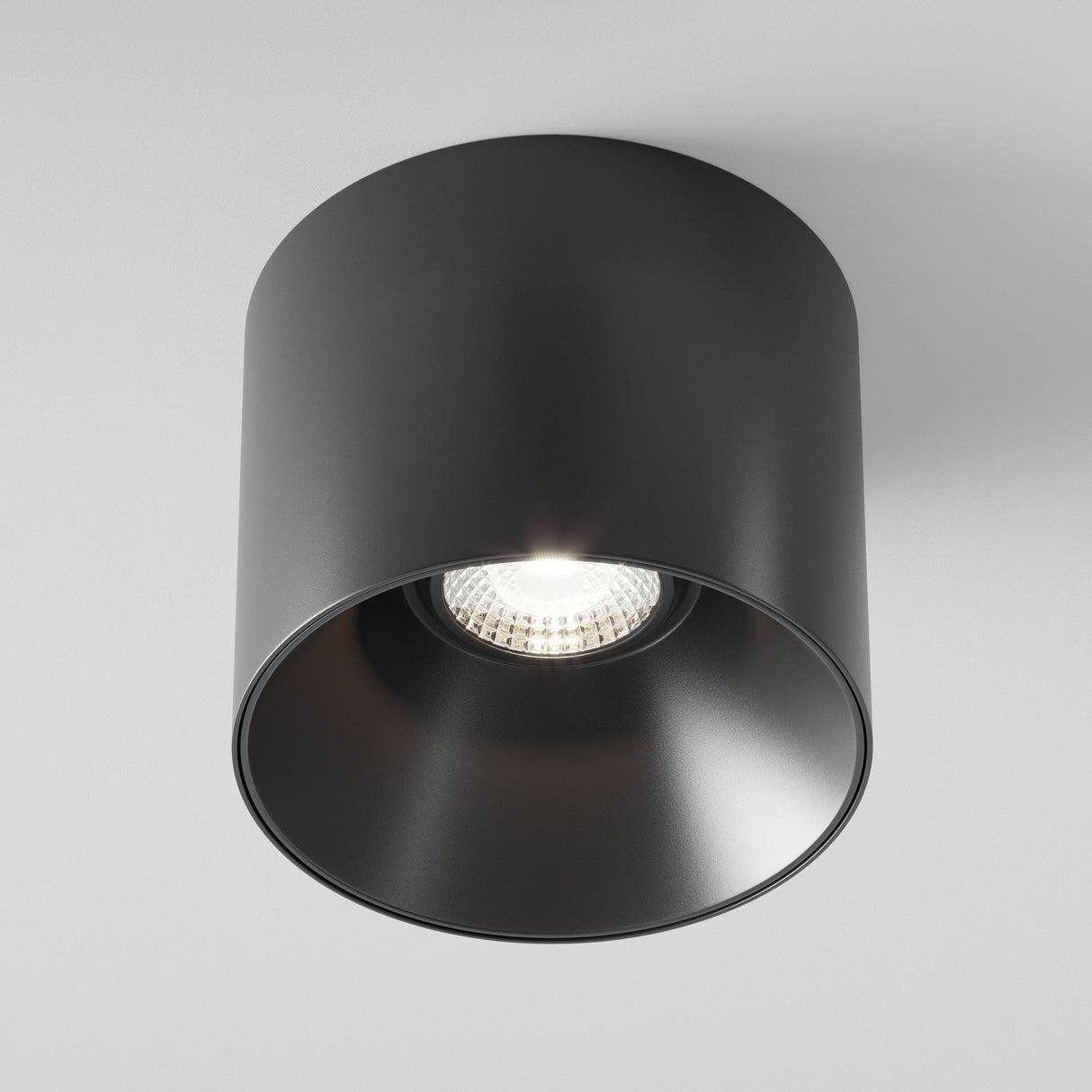 The Alfa LED Dimmable Fixed Round Surface Downlight 15W 4000K in black is elegantly mounted on a white ceiling. It features a sleek cylindrical design with a visible bulb, emitting cool white light downward for an inviting ambiance.