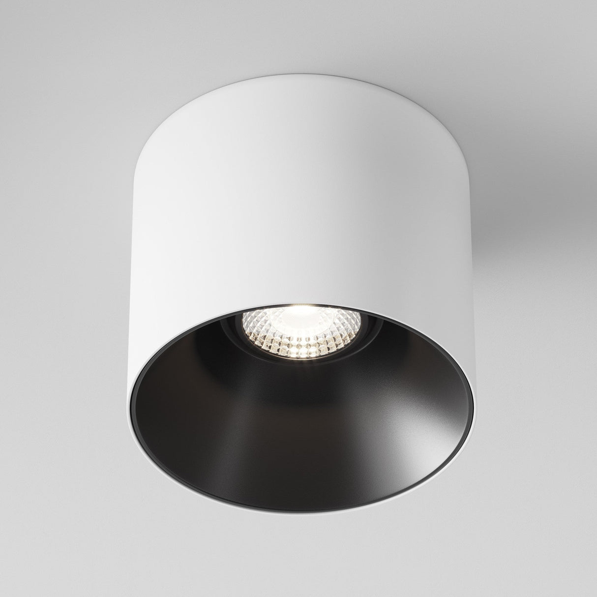 The Alfa LED Fixed Round Surface Downlight 12W 4000K features a cylindrical white exterior and black interior. The illuminated cool white bulb emits a subtle glow, ideal for enhancing contemporary spaces and is mounted on a white ceiling.