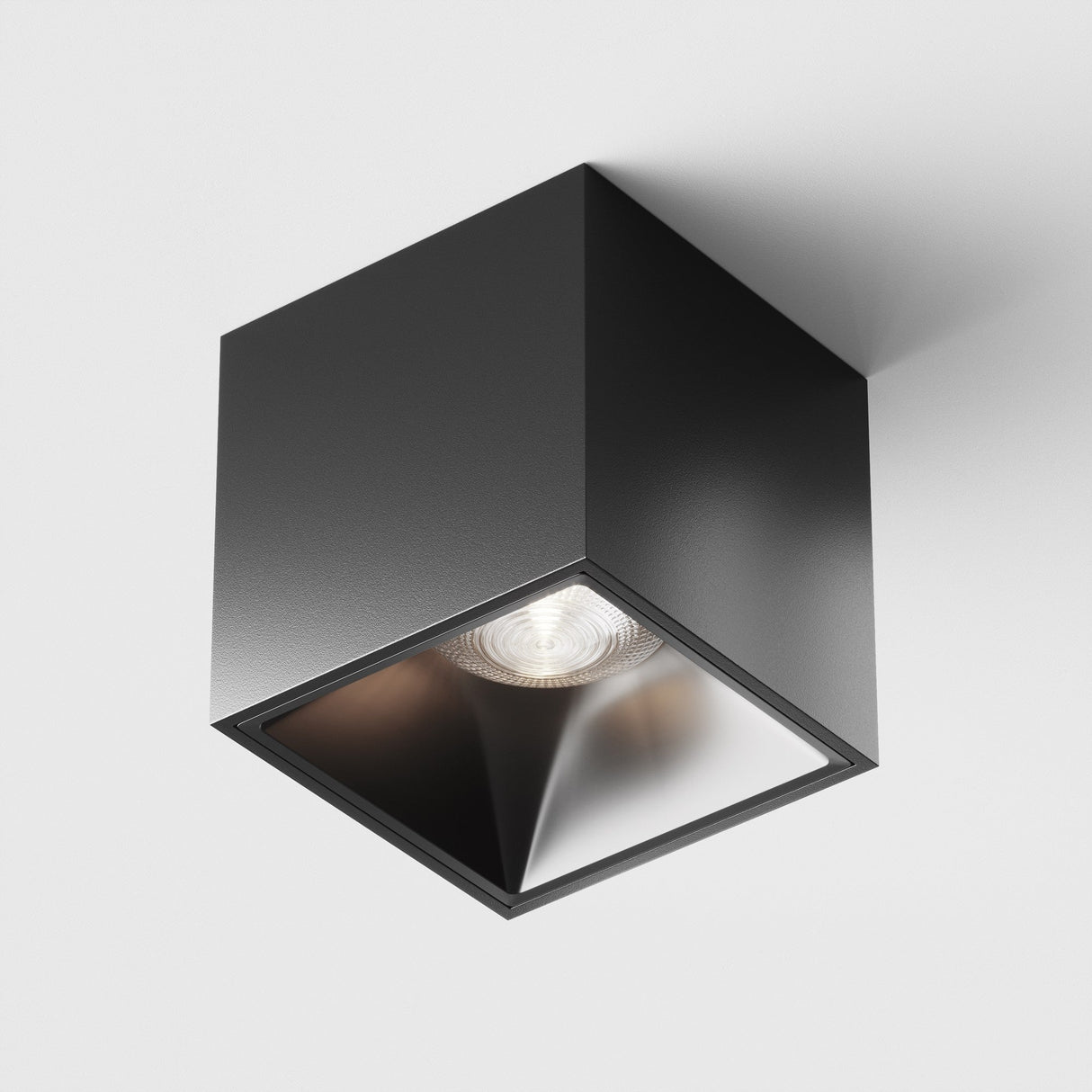 The Alfa LED Square Surface Downlight 12W 4000K in black is a modern cube-shaped ceiling light fixture with a recessed cool white LED, offering bright illumination. Its sleek black finish stands out against a white background, highlighting its geometric shape for a minimalist statement.