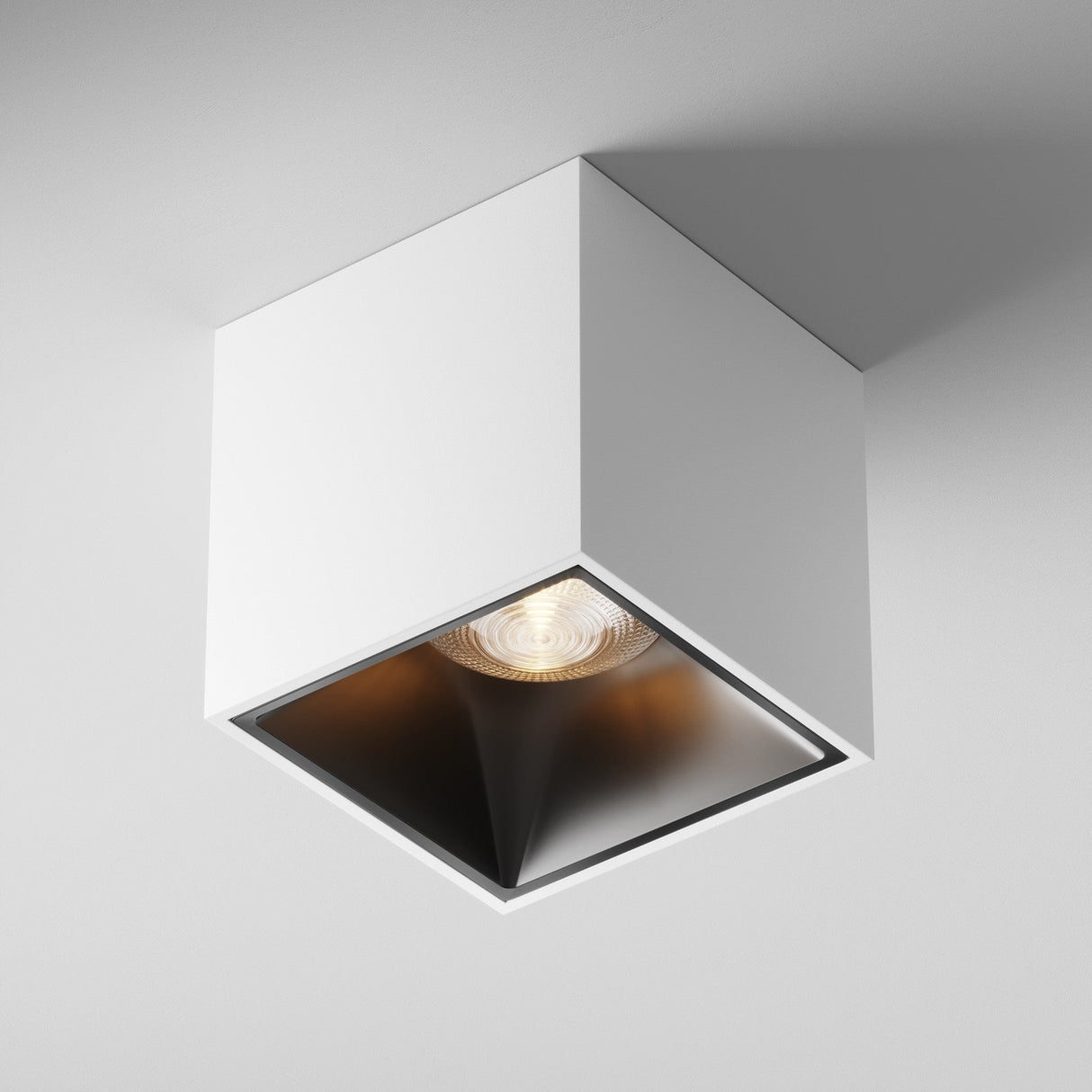 The Alfa LED Square Surface Downlight 12W 3000K features a modern, white cube design emitting a focused beam from a recessed bulb. Its dark interior elegantly contrasts with the bright exterior.