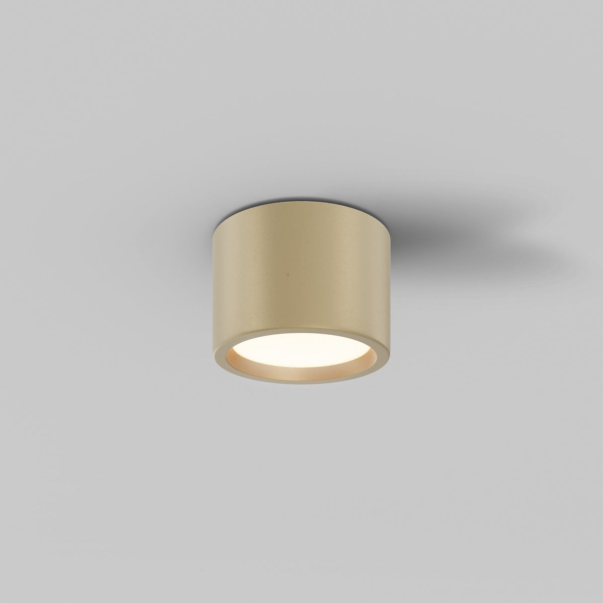 The Hydra LED Round Surface Downlight 10W 3000K with a matte gold finish is mounted on a gray ceiling, featuring a cylindrical design and IP44 rating. It emits a warm glow downward, casting subtle shadows.