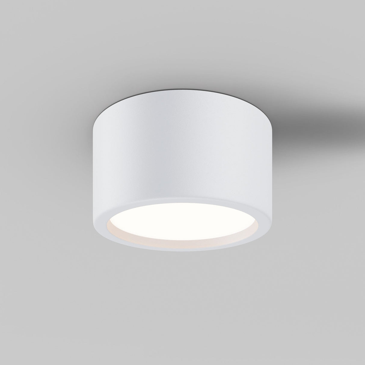The Hydra LED Round Surface Downlight 15W 3000K in white, IP44 rated, is a modern cylindrical fixture on a light gray ceiling, emitting a soft glow that casts subtle shadows around its edges.
