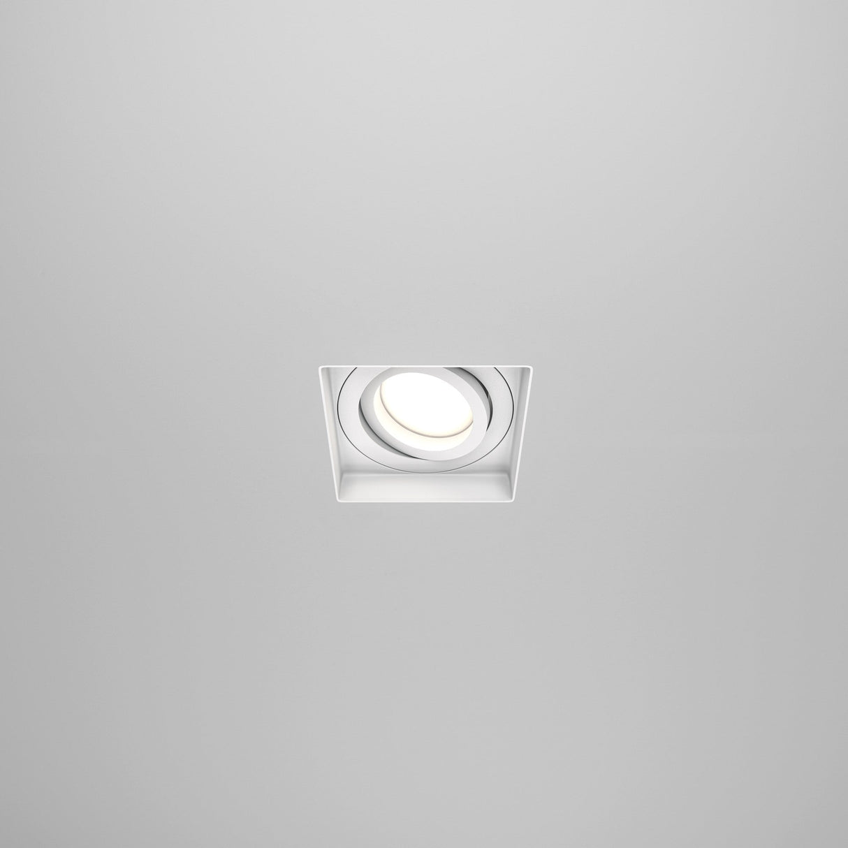 The Atom Single Tilt Square Plaster-in Recessed Downlight - White features a sleek square design and integrates seamlessly into light-colored ceilings, emitting a bright white glow for a minimalist, modern look.