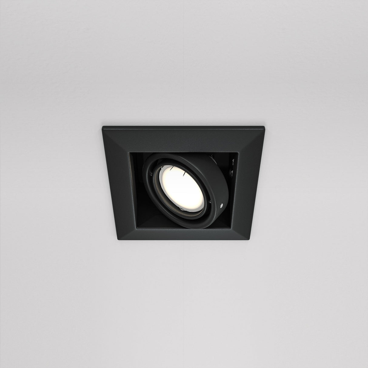 The Metal Modern Single Tilt Square Recessed Downlight in black features a sleek square frame, with an adjustable circular bulb set at an angle. This contemporary fixture includes a swivel mechanism, casting soft white light against a white ceiling.