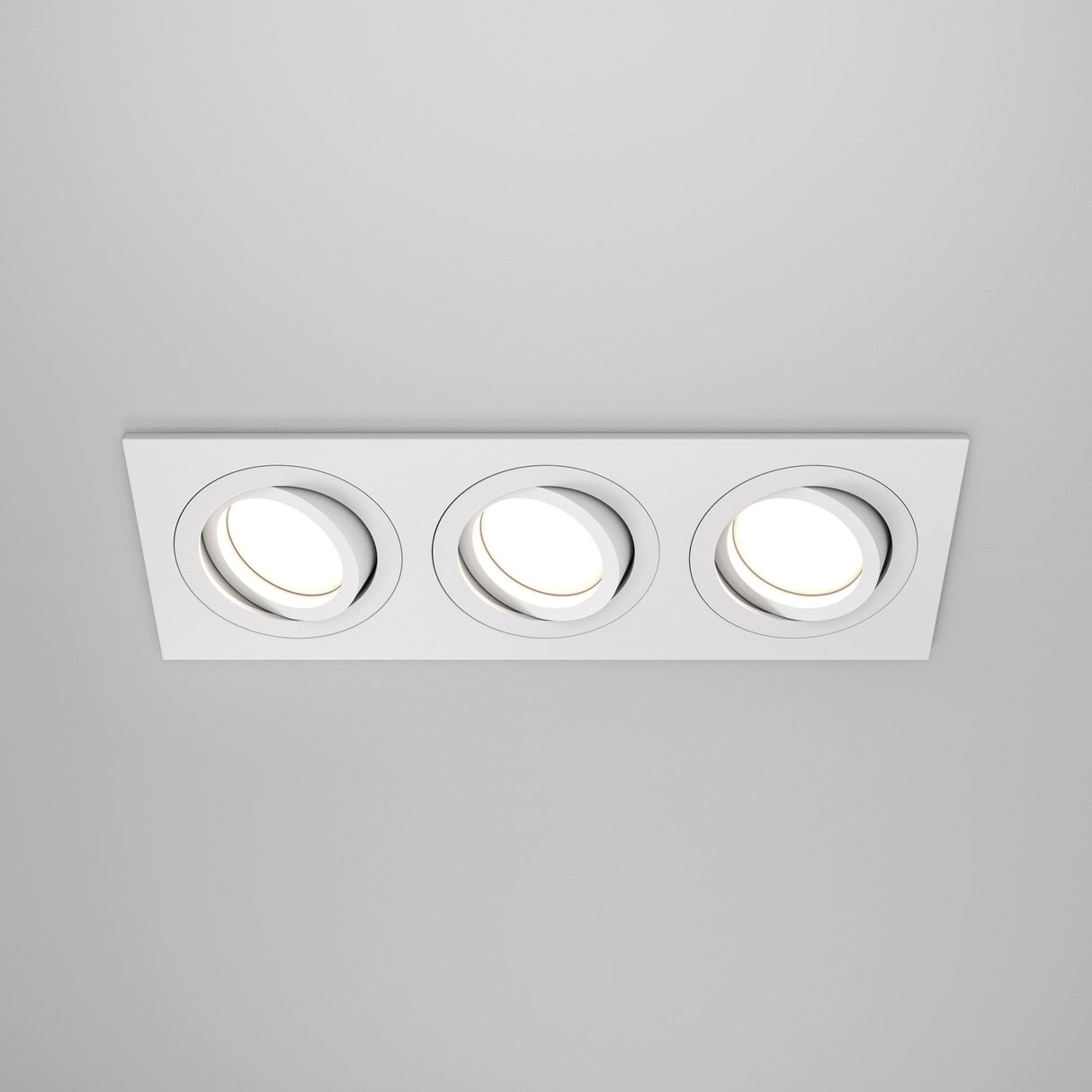 The Atom Tri Tilt Rectangular Recessed Downlight - White features a modern design with three recessed circular lights, white trim, and adjustable tilt for customized angles. Set against a gray ceiling, the lights create a bright glow when turned on.