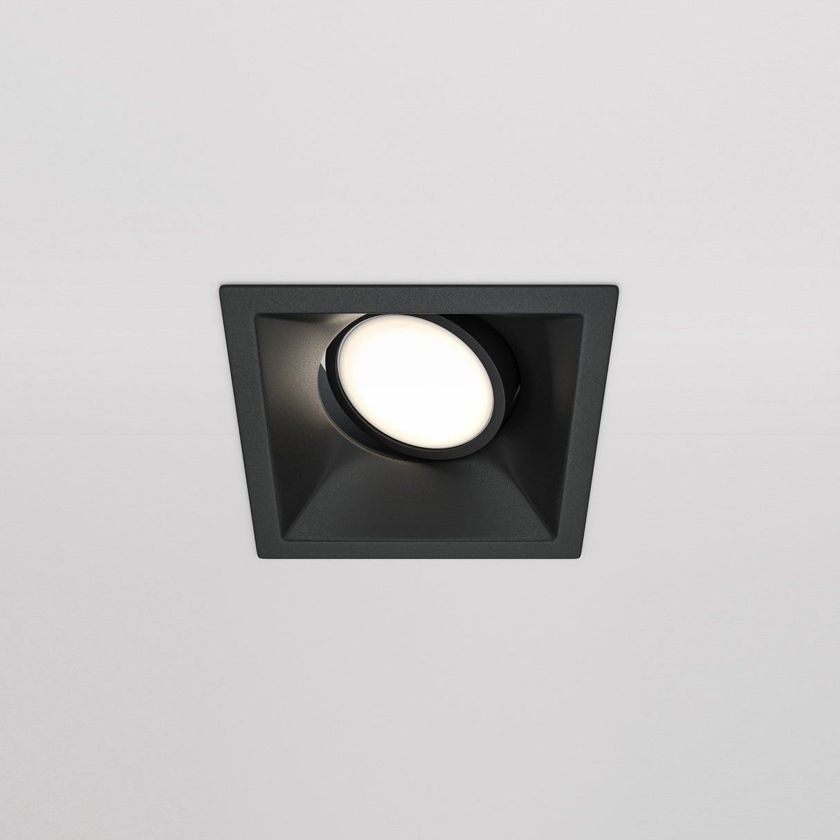 The Dot Single Tilt Square Recessed Downlight - Black is a minimalist lighting fixture with a circular LED bulb in a square design, providing a soft glow against a white ceiling.